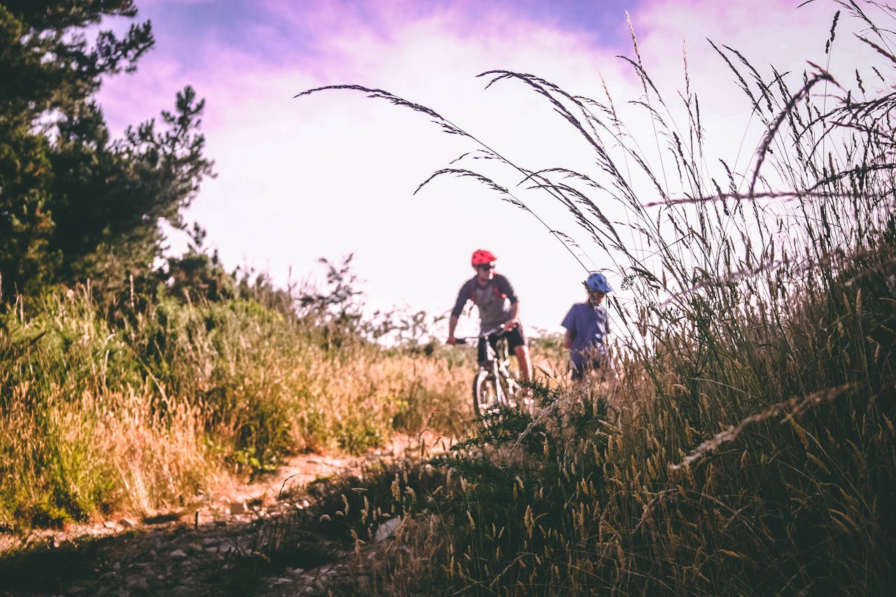 Two cyclists wearing helmets navigate through a scenic unpaved path surrounded by tall grass and natural landscape.