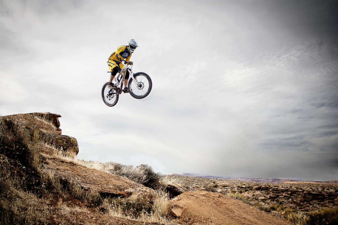 A cyclist performs a daring jump off a rocky cliff under a cloudy sky.